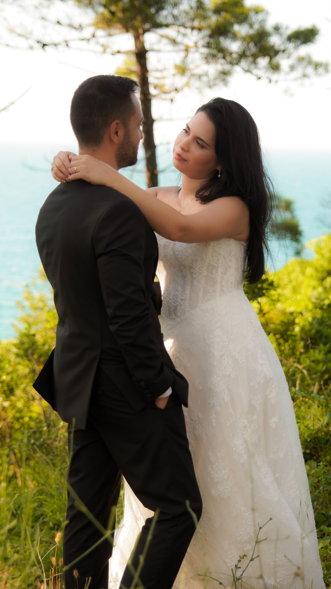 Sposa in abito di pizzo che abbraccia lo sposo in abito scuro, ritratto di coppia tra il verde con il mare in lontananza