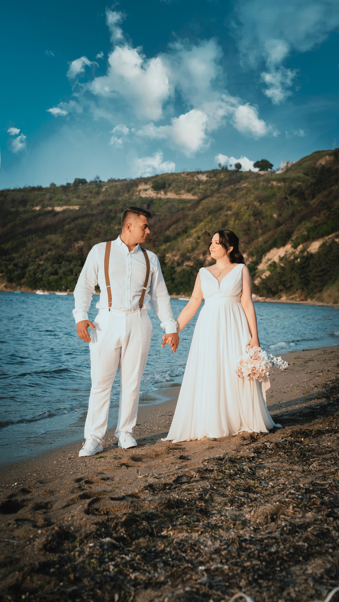 Sposi a piedi nudi sulla sabbia che camminano tenendosi per mano, lei con bouquet pastello e mare sullo sfondo